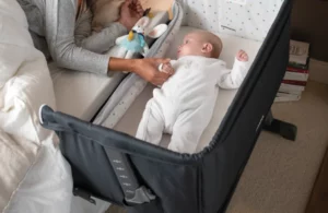 Un bébé est allongé dans son lit de cododo, tout près de sa mère, qui pose sa main sur lui